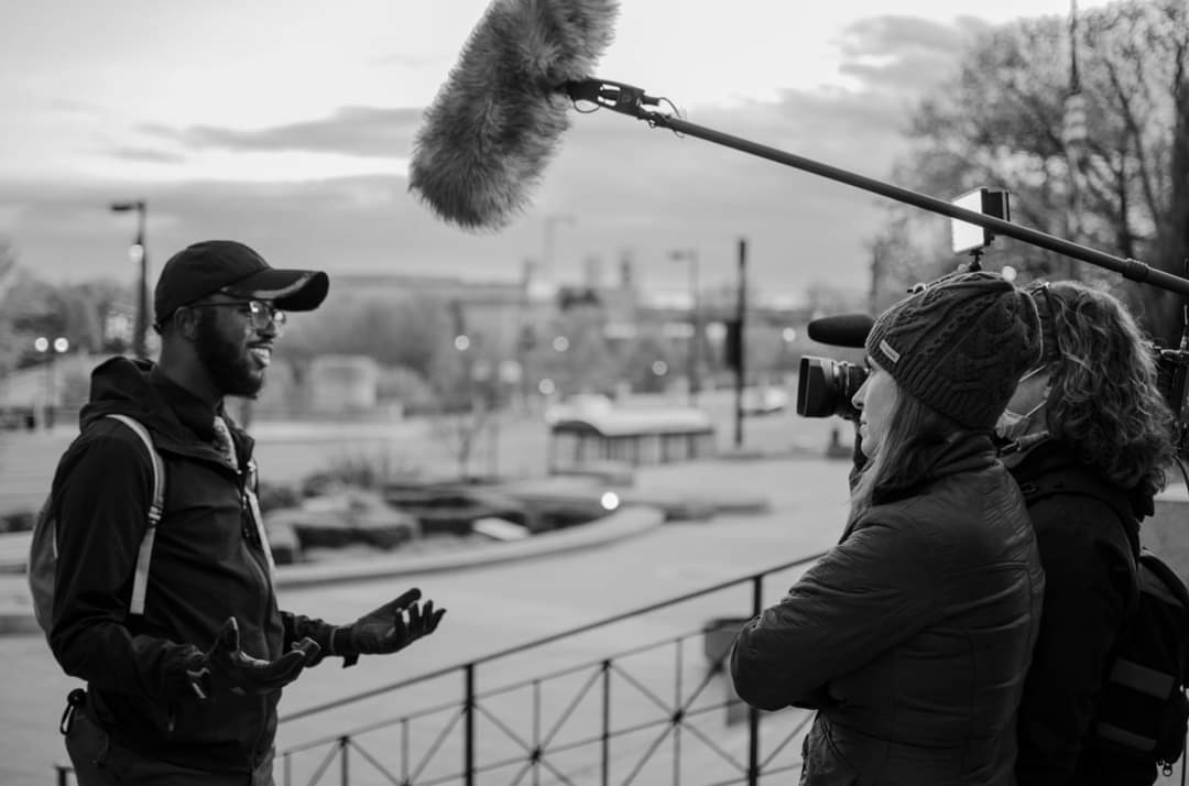 Black and white frame of DaVante' Goins speaking to a camera crew